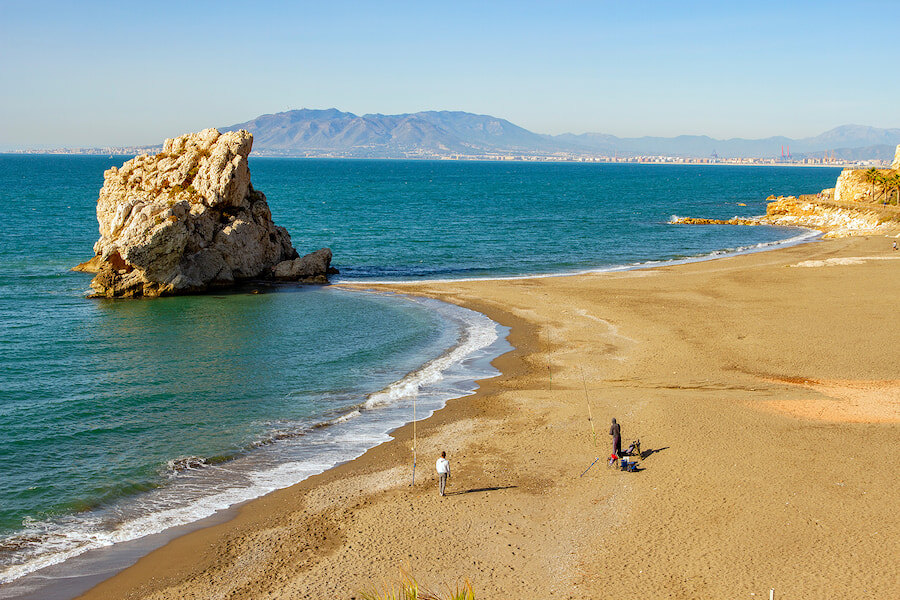 Fischer an einem ruhigen Sandstrand in Málaga mit einer großen Felsformation im Meer, ruhigem blauen Wasser und in der Ferne unter einem klaren Himmel sichtbaren Bergen.