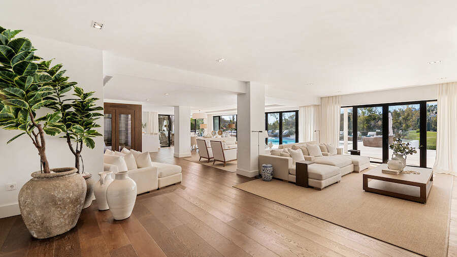 Amplia y luminosa sala de estar contemporánea en una villa de lujo en Marbella, con suelos de madera natural, grandes puertas corredizas de vidrio que se abren a la terraza y la piscina y una elegante decoración en tonos neutros.