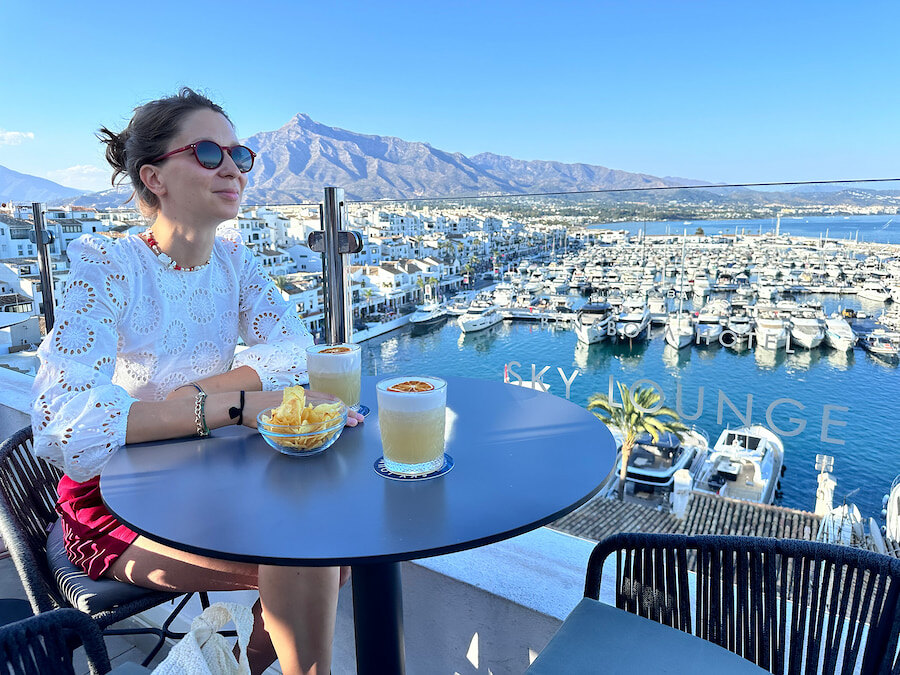 Woman enjoying a drink at Sky Lounge | Benabola, overlooking the Puerto Banús marina in Marbella. The rooftop setting offers spectacular views of luxury yachts, white buildings, and the La Concha mountain in the background on a clear sunny day.