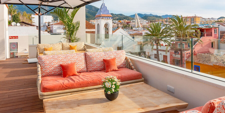 Vibrant rooftop terrace at Sky Bar | La Fonda Heritage Hotel Relais & Châteaux, featuring colourful cushioned seating, wooden tables, and city views with traditional rooftops, palm trees, and mountain peaks in the background. A blue-and-white tiled dome and modern bridge add local charm to the scene.