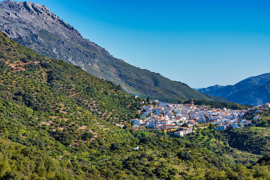 Charming whitewashed village nestled among lush green hills and terraced fields, set against the backdrop of towering mountains under a bright blue sky.