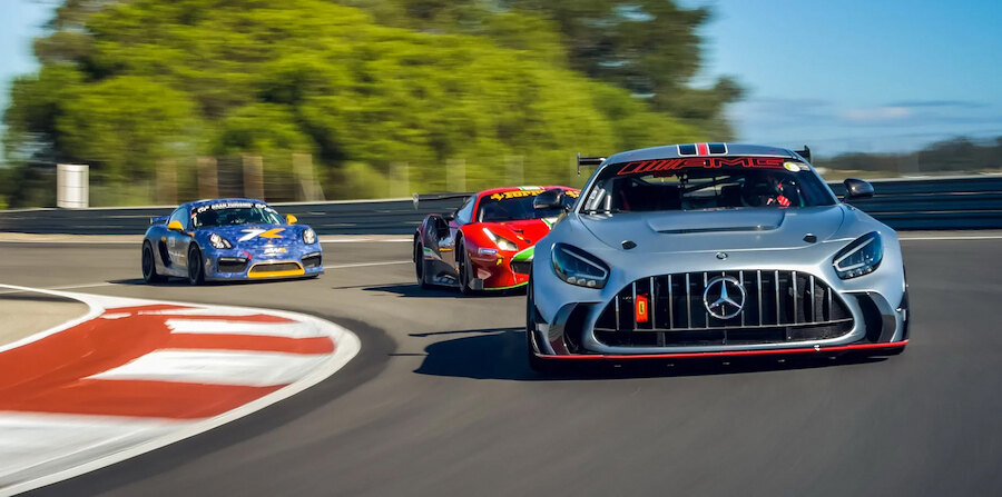 Racetracks near Marbella. Three high-performance race cars—led by a silver Mercedes-AMG—speed through a racetrack corner during a competitive session, with a Porsche and Ferrari closely following, set against a backdrop of trees and clear blue sky.