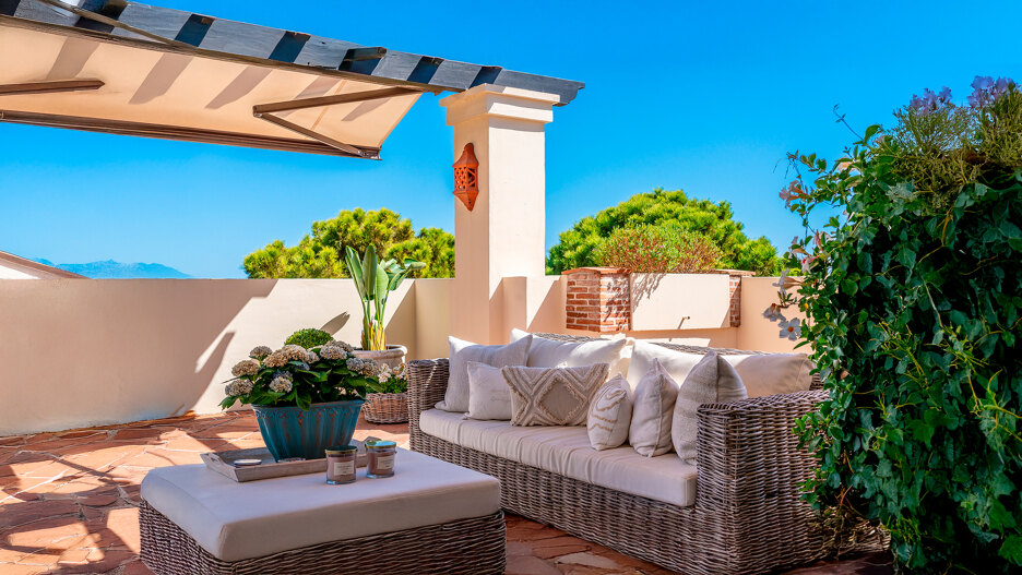 Terraza soleada con sofá de mimbre, cojines, plantas en macetas y vistas a la montaña bajo un cielo azul.
