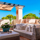 Sunny terrace with wicker sofa, cushions, potted plants and mountain views under a blue sky.