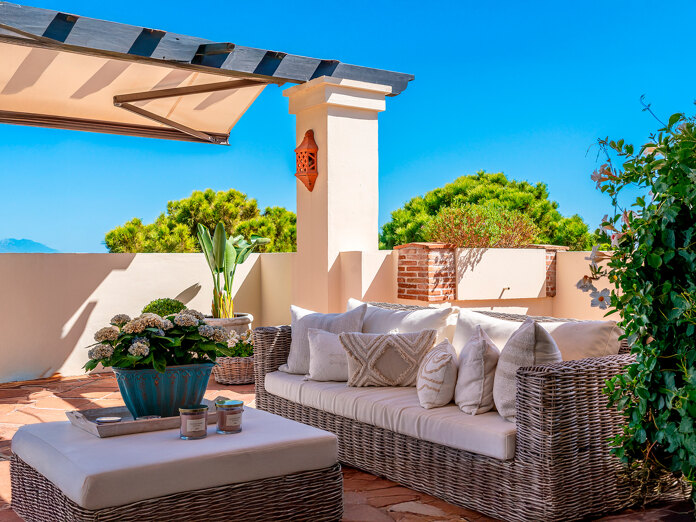 Sunny terrace with wicker sofa, cushions, potted plants and mountain views under a blue sky.