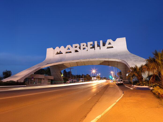 Iconic Marbella entrance arch illuminated at dusk, with traffic lights creating motion trails on the main coastal road