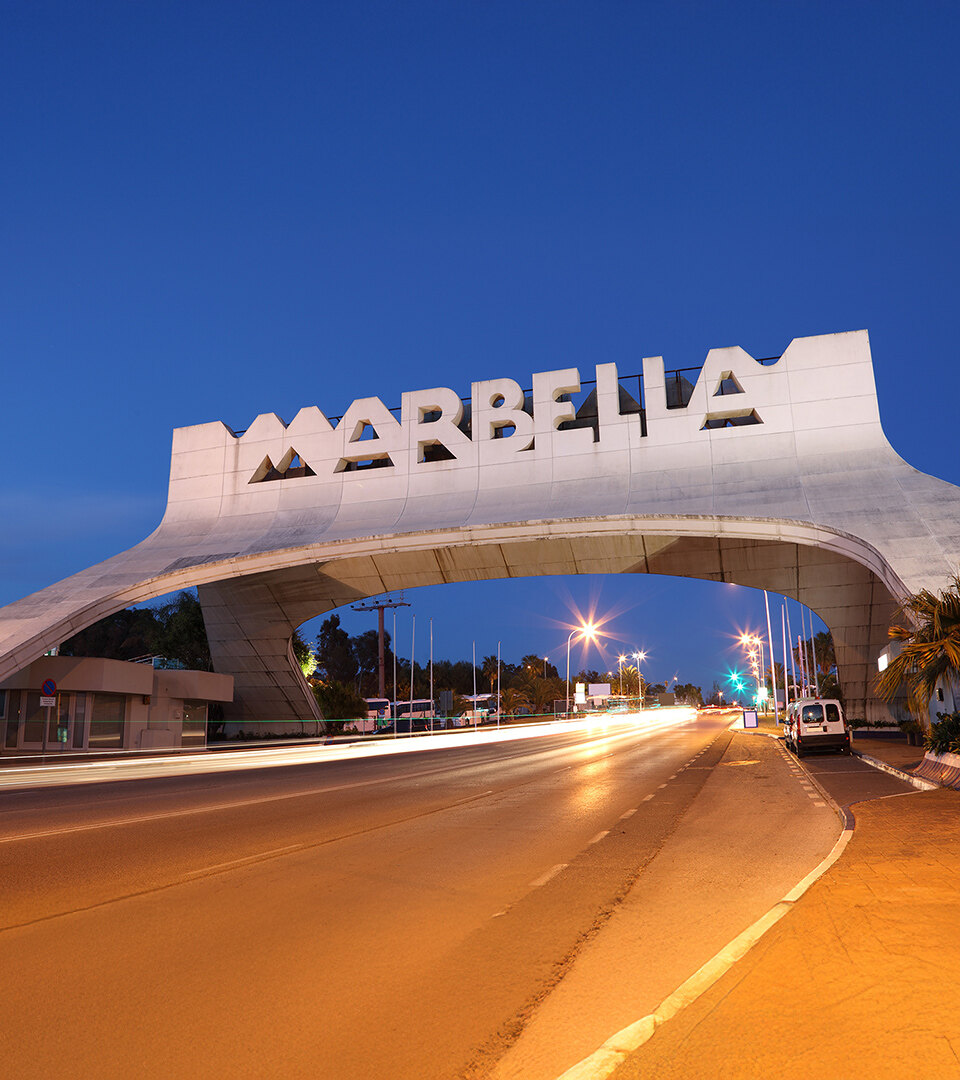 Iconic Marbella entrance arch illuminated at dusk, with traffic lights creating motion trails on the main coastal road