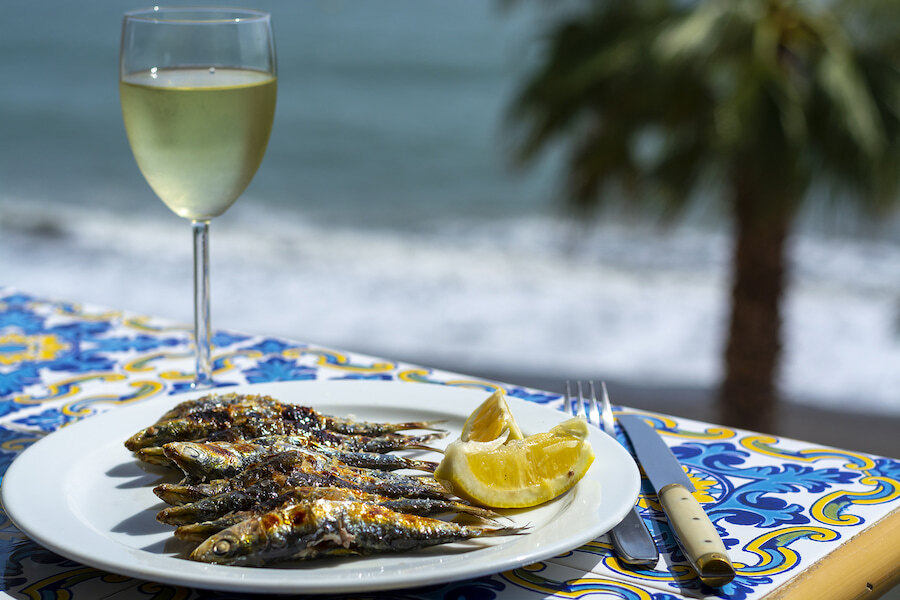 Plate of grilled sardines with lemon wedge and a glass of white wine, served on a colourful tiled table by the seaside in Marbella