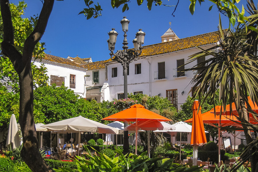JamesEdition's Top Lifestyle Cities. Sunny courtyard in Marbella’s Old Town with vibrant orange and white parasols, lush greenery, and traditional Andalusian architecture in the background