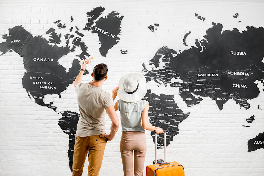 Couple avec une valise regardant une grande carte du monde accrochée au mur, pointant vers différents pays.