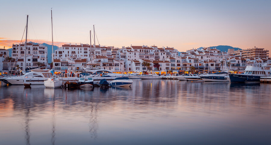 Jachthaven van Puerto Banús bij zonsondergang met jachten, kalm water en witte mediterrane gebouwen.