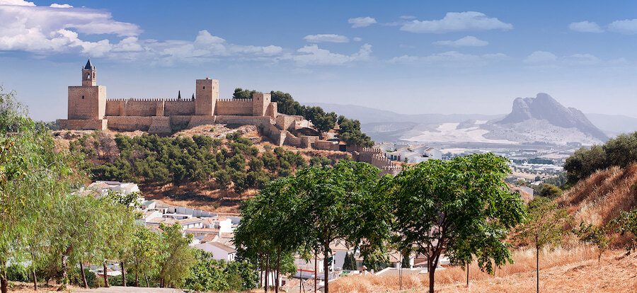 Locatie: Antequera, ongeveer 1 uur rijden ten noorden van Marbella. Openingstijden: Het hele jaar door; gratis toegang tot de meeste monumenten. Bezoekerscentra: Begin in het Dolmenmuseum in Antequera of in het bezoekerscentrum van El Torcal. Panoramisch uitzicht op het Alcazaba van Antequera, Málaga, een Arabisch fort gelegen op een heuvel, omringd door bomen en witgekalkte huizen, met de berg Peña de los Enamorados op de achtergrond onder een helderblauwe lucht.