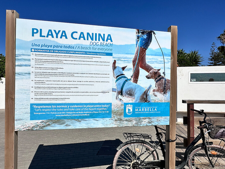 Guide de Marbella pour chiens. Panneau d’information sur la plage dog-friendly de Marbella indiquant les règles pour les chiens, avec une image de deux chiens jouant dans l’eau avec leur maître.