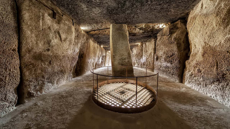 Werelderfgoed. Interieur van de dolmen van Menga in Antequera, Málaga, met de karakteristieke centrale stenen pilaar, gebeeldhouwde rotswanden, subtiele plafondverlichting en een beschermd metalen rooster op de vloer.