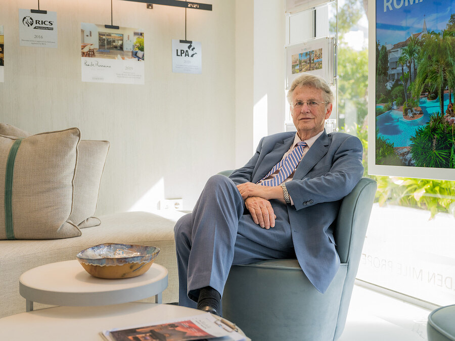 Christopher Clover, Geschäftsführer von Panorama Properties, sitzt in einem modernen Sessel, trägt einen blauen Anzug und eine gestreifte Krawatte in einem hellen Büro. Neben ihm stehen Beistelltische mit Zeitschriften und einer dekorativen Schale, während an der Wand und im Fenster Poster und Fotos von Luxusimmobilien hängen.