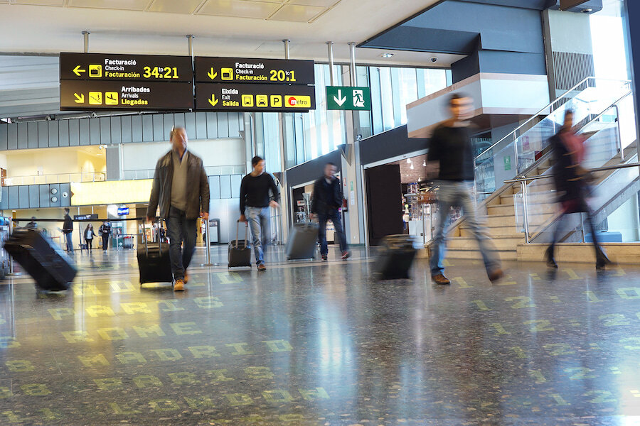 Passengers walking with rolling suitcases inside Málaga Airport terminal, with yellow directional signs overhead.