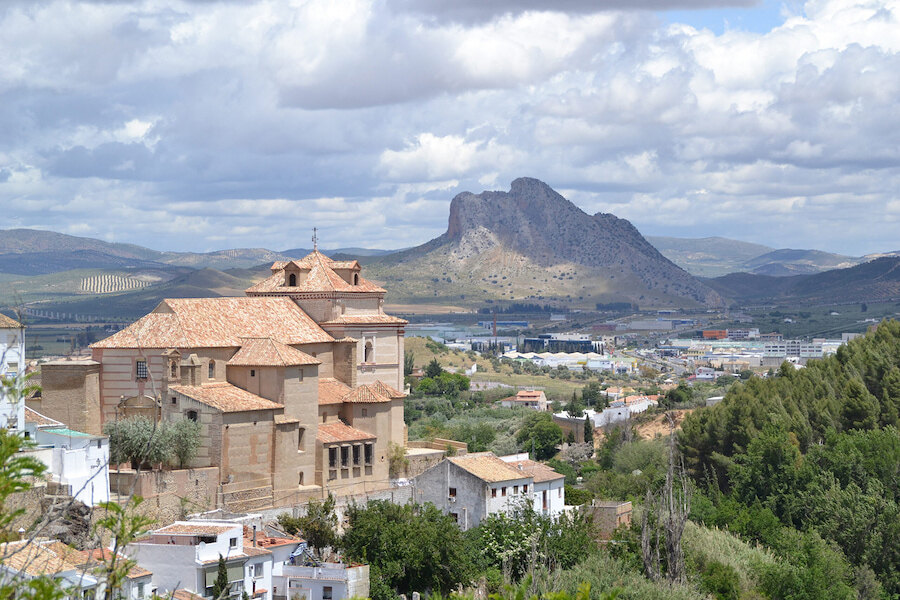 Panoramisch uitzicht op Antequera, Málaga, met de prominente berg Peña de los Enamorados op de achtergrond en de historische Koninklijke Collegiale Kerk van Santa María la Mayor op de voorgrond, omringd door witgekalkte huizen en groene heuvels onder een gedeeltelijk bewolkte hemel