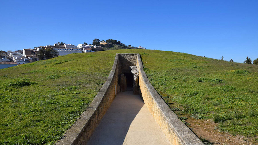Ingang van de Viera Dolmen in Antequera, Málaga, met een stenen gang die naar een met gras bedekte heuvel leidt, onder een helderblauwe lucht met de stad zichtbaar op de achtergrond.