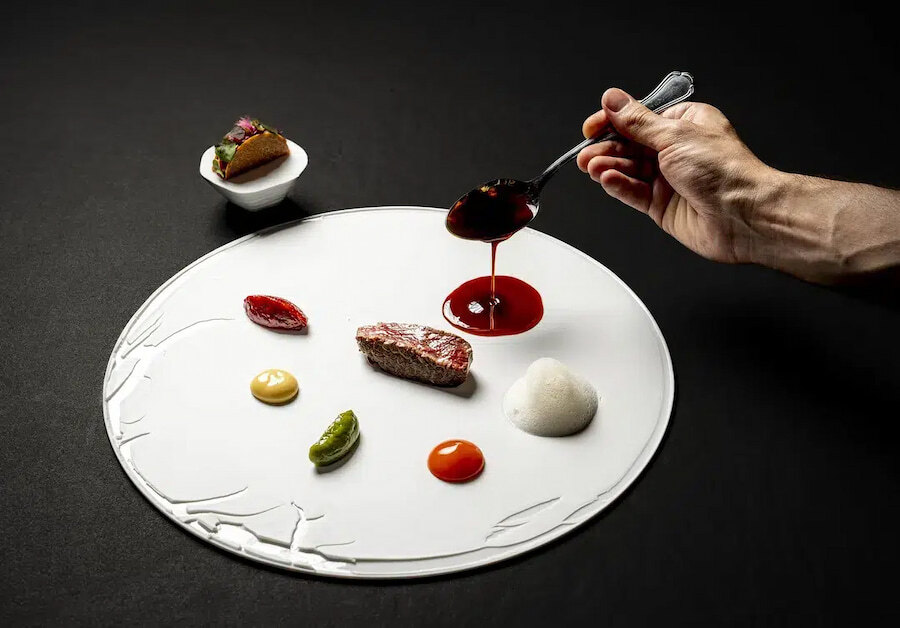 Marbella's Michelin-Star restaurants: Fine Dining Experiences in Marbella. A haute cuisine dish, carefully presented on an elegant white base. The plate features a piece of meat accompanied by vibrantly colored sauces such as red, yellow, and green, along with a white foam. A hand is pouring additional sauce from a spoon, adding a final touch of detail to the composition. In the upper left corner, a small container with a decorative touch can be seen. The dark background highlights the sophistication and delicacy of the dish, reflecting a luxurious dining experience.