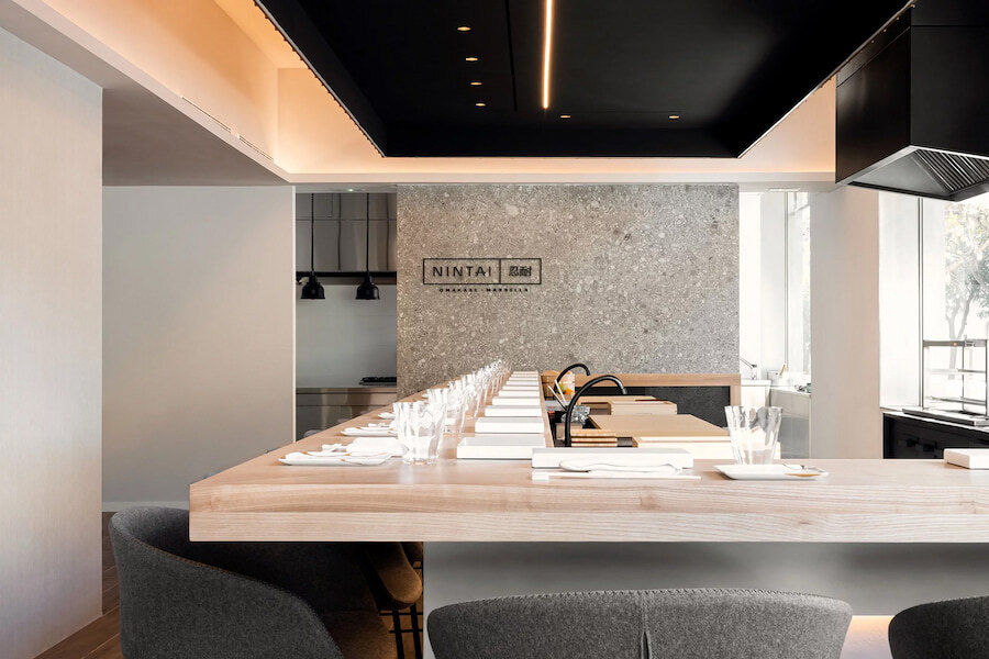 Nintia Restaurant. The image shows a modern and elegant restaurant with a light wood bar and gray chairs. The 