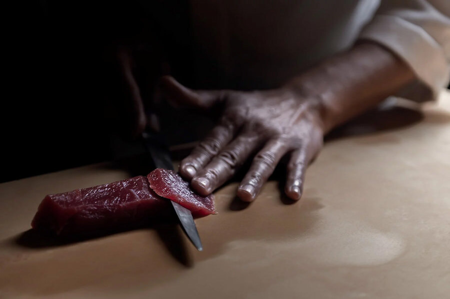 Nintia Restaurant. The image shows a chef carefully cutting a piece of fresh tuna with a knife, while his hand holds the fish. The dim lighting highlights the precision and skill in the preparation, reflecting the attention to detail and skill in the kitchen.