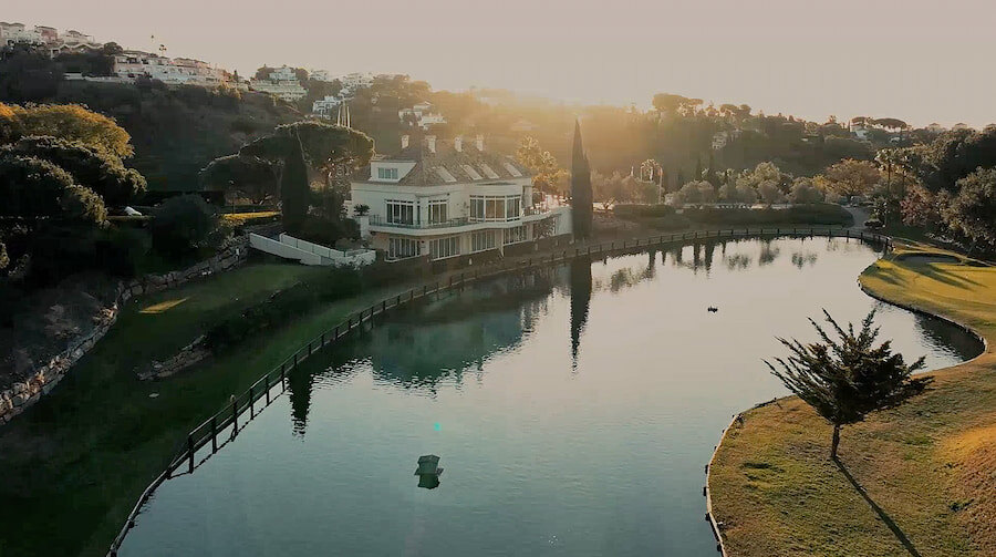 El Lago Restaurant: The image shows an aerial view of an elegant lakeside restaurant, surrounded by gardens and trees. The soft sunset light creates a serene and relaxing atmosphere, highlighting the natural and architectural beauty of the space.