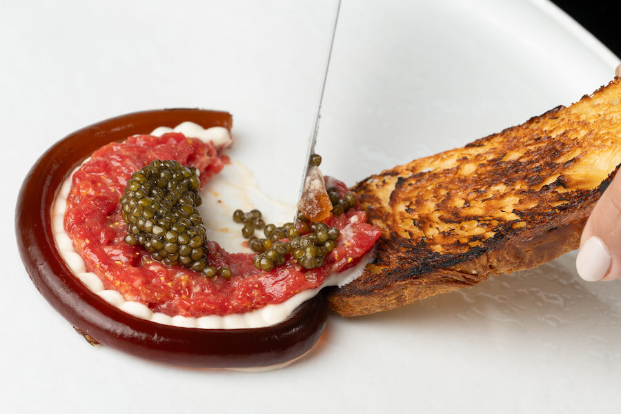 Back Restaurant: The image shows a delicious gastronomic preparation with caviar and a mix of ingredients on a gelatinous base, accompanied by golden toast. A knife gently cuts the dish, highlighting its texture and detail.