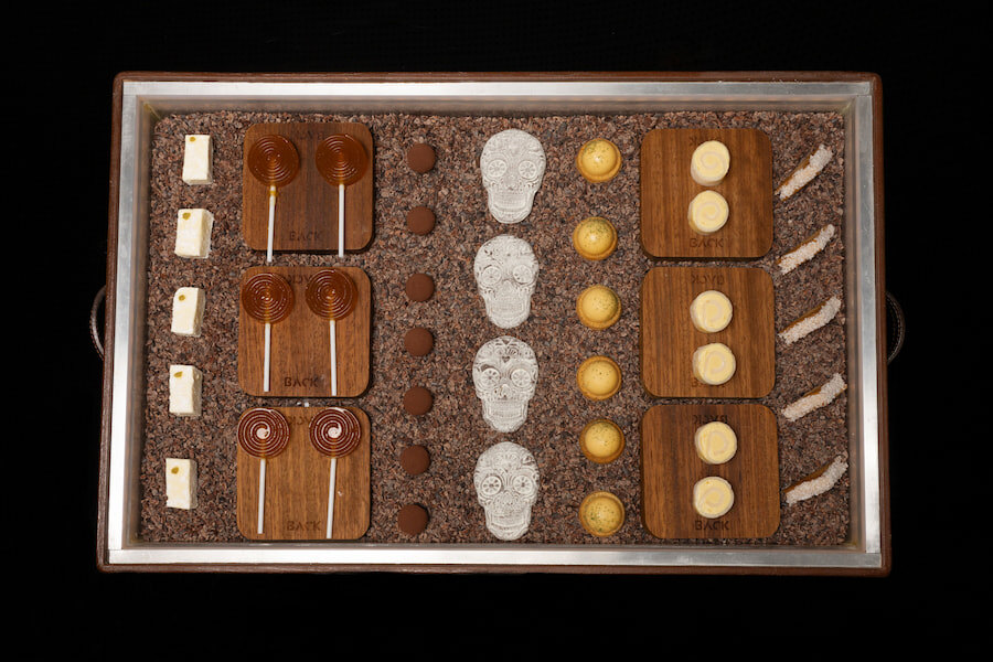 Back Restaurant: The image shows an elegant tray with a variety of fine sweets arranged neatly. It includes swirly lollipops, chocolates, macaroons, and decorative skull-shaped figurines. Everything is presented on a cocoa bean base, creating an attractive and sophisticated visual contrast.