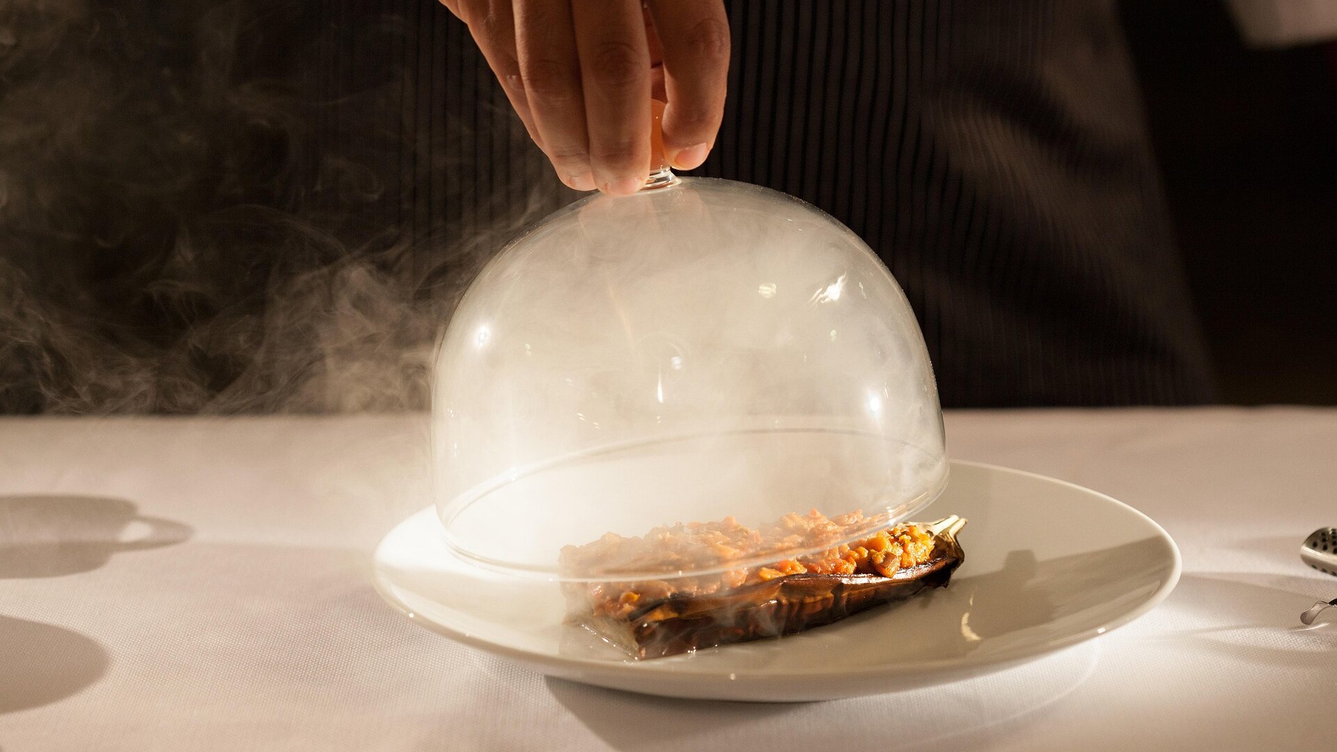 Gourmet dish served under a glass cloche with smoke, being uncovered by a waiter’s hand.