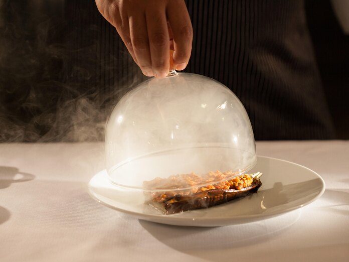 Gourmet dish served under a glass cloche with smoke, being uncovered by a waiter’s hand.
